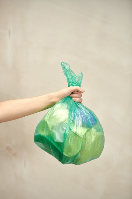 A human hand grasping a loosely knotted green plastic rubbish bag filled with waste, held against a plain, neutral background. The bag appears semi-transparent with hints of blue and yellow tones visible through the material. The hand is positioned in the foreground, with fingers wrapping around the bag's gathered top, which is twisted and tied. The scene suggests an act of waste disposal or rubbish collection, typically relevant to private waste management and alternative rubbish removal services. There are no other objects or environmental elements visible, focusing solely on the hand and the rubbish bag, emphasizing the process of rubbish handling in a neutral, professional manner consistent with waste removal service contexts.