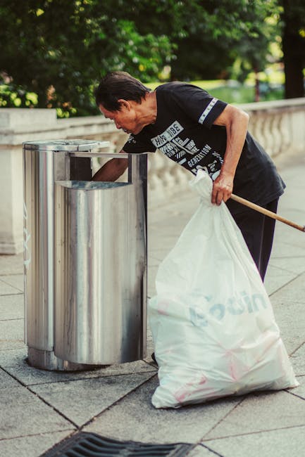 A middle-aged man with dark hair wearing a black t-shirt with printed text and dark trousers is seen disposing of rubbish into a tall, cylindrical, stainless steel recycling bin on a paved sidewalk. The bin features a wide, rectangular opening at the top with a smooth, metallic finish that reflects the surrounding environment. The man is bending slightly forward, holding a white plastic bag filled with waste in his left hand, while his right hand is reaching into the bin to discard additional rubbish. Behind him, there is a low stone barrier and lush green trees, suggesting an outdoor park or urban green space setting. The scene is illuminated by natural daylight, casting soft shadows on the pavement. This capture highlights the process of individual waste disposal, which may relate to private rubbish handling or independent collection services, aligning with the themes of rubbish removal and recycling support provided by Rubbish Removal Stockwell.