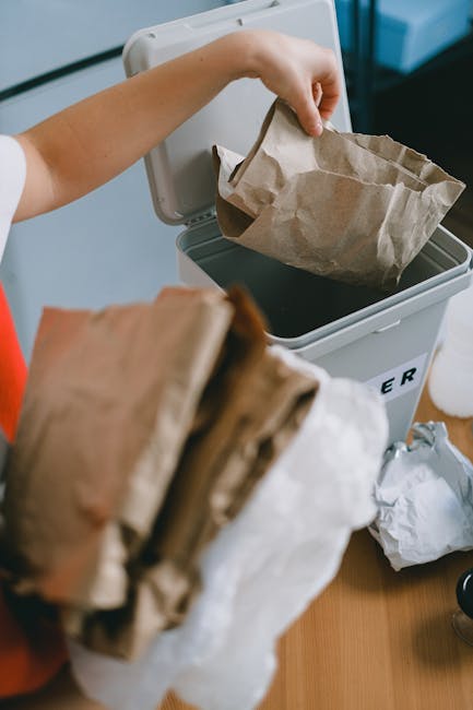 A person is seen disposing of brown paper waste bags into a grey recycling bin with the lid open, situated on a wooden surface indoors. The person's arm is visible, wearing a white sleeve, and holding one of the paper bags over the bin. Several crumpled paper bags are also held in their hand, ready for collection. Surrounding the bin are additional paper waste items, including crumpled and folded bags, some partially on the surface and some on the floor nearby. The environment appears to be a domestic or office setting with a focus on waste segregation. The scene depicts an aspect of private waste disposal or rubbish separation, which relates to alternative rubbish removal methods, highlighting the importance of proper refuse management advocated by companies like Rubbish Removal Stockwell in the context of recycling and waste collection services.