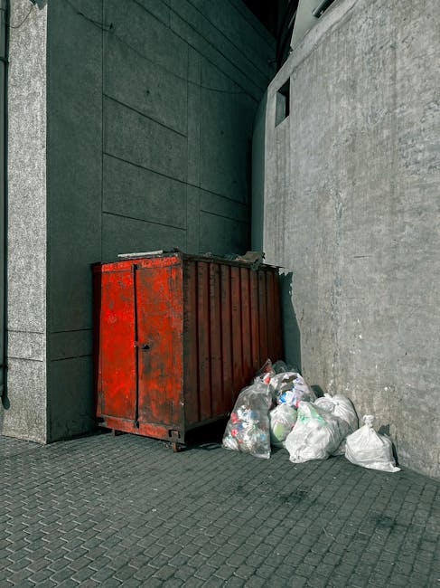 The image depicts a red metal waste container positioned against a dark green wall on the left and a light grey textured wall on the right, within an outdoor urban setting. The container, which has a weathered and slightly rusted finish, is large and rectangular, with a flat top and vertical ridges along its sides. Several clear plastic bags filled with mixed waste materials are placed on the ground beside it, some leaning against the container. The bags contain various discarded items, with visible paper, plastic bottles, and other waste materials. The ground surface is paved with dark grey interlocking bricks, and natural daylight illuminates the scene, casting subtle shadows. The surrounding environment suggests an alleyway or rear access point, typical for independent waste collection or private rubbish disposal in a commercial or residential area, with no visible signage or markings indicating municipal collection services. The overall scene reflects a typical scene for private rubbish removal services such as those provided by Rubbish Removal Stockwell, serving local waste management needs.