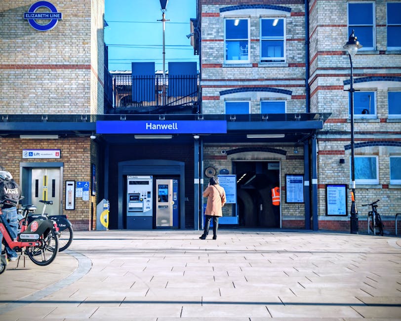 A woman stands in front of Hanwell station entrance, which is part of an older brick building with a mix of cream and red bricks, and three large multi-pane windows on the upper floors. The station features a dark blue overhang with clear signage indicating 'Hanwell' and the Elizabeth Line logo in the top left corner. In the foreground, there are two bicycles parked on the left, one with a black and red basket, and a yellow contactless payment terminal for the lift on the wall. To the right of the station entrance, an orange safety vest is visible on a person inside the entrance, and there are several information boards and notices mounted on the wall. A street lamp with a black post and lantern-style light is situated on the right side of the building, and the pavement consists of large, light-colored rectangular tiles. The scene is set during daylight with clear, natural lighting, and the overall environment suggests a busy urban area with facilities for public transport and bicycle access, relevant to private or alternative waste handling in the vicinity managed by local services like Rubbish Removal Stockwell.