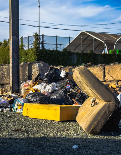 The image depicts a red metal waste container positioned against a dark green wall on the left and a light grey textured wall on the right, within an outdoor urban setting. The container, which has a weathered and slightly rusted finish, is large and rectangular, with a flat top and vertical ridges along its sides. Several clear plastic bags filled with mixed waste materials are placed on the ground beside it, some leaning against the container. The bags contain various discarded items, with visible paper, plastic bottles, and other waste materials. The ground surface is paved with dark grey interlocking bricks, and natural daylight illuminates the scene, casting subtle shadows. The surrounding environment suggests an alleyway or rear access point, typical for independent waste collection or private rubbish disposal in a commercial or residential area, with no visible signage or markings indicating municipal collection services. The overall scene reflects a typical scene for private rubbish removal services such as those provided by Rubbish Removal Stockwell, serving local waste management needs.